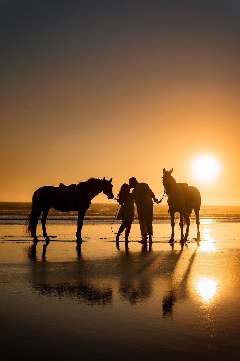 Couple with two horses on the beach, silhouetted at sunset