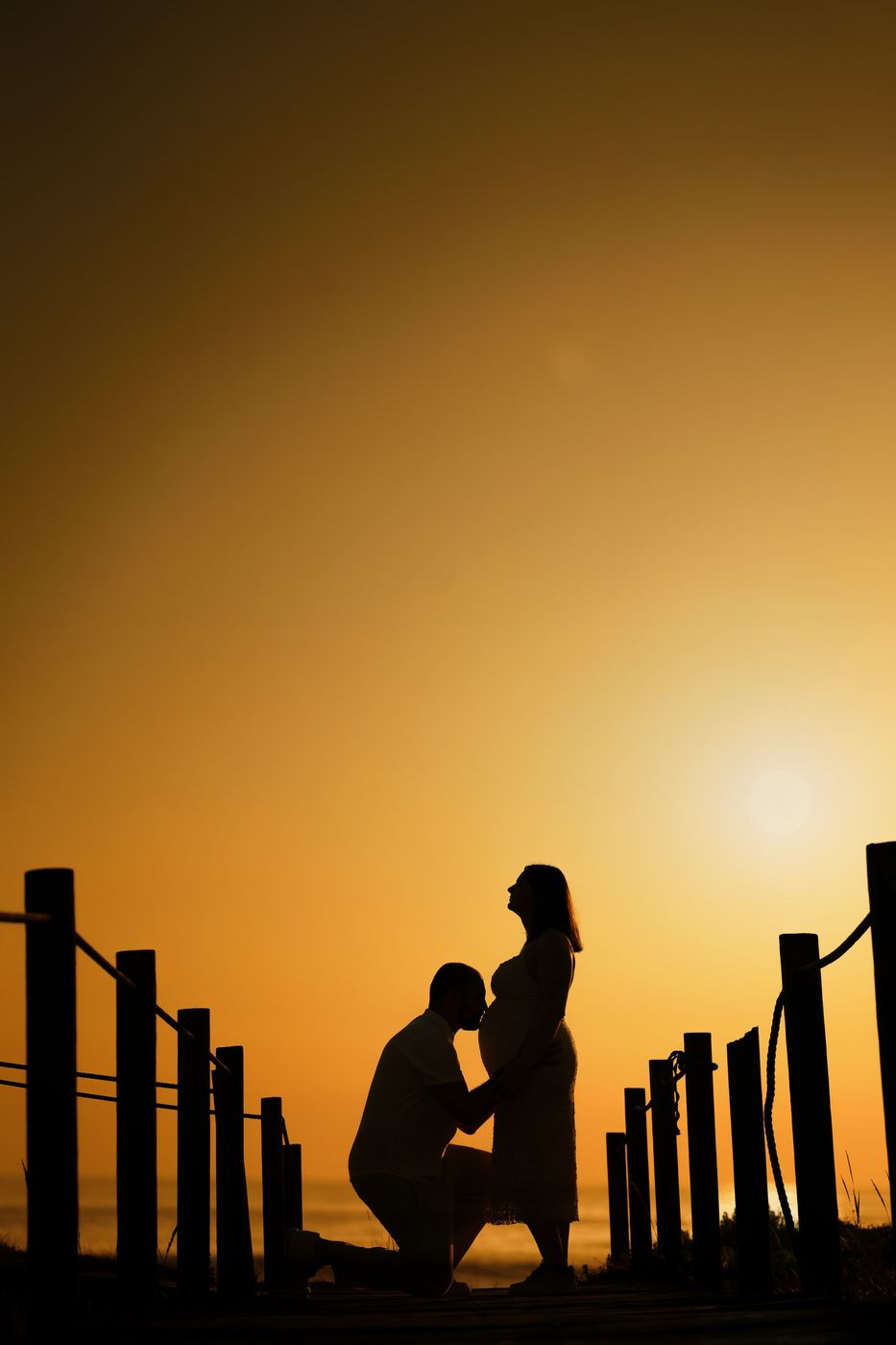 Couple silhouetted at sunset, him kissing the bump