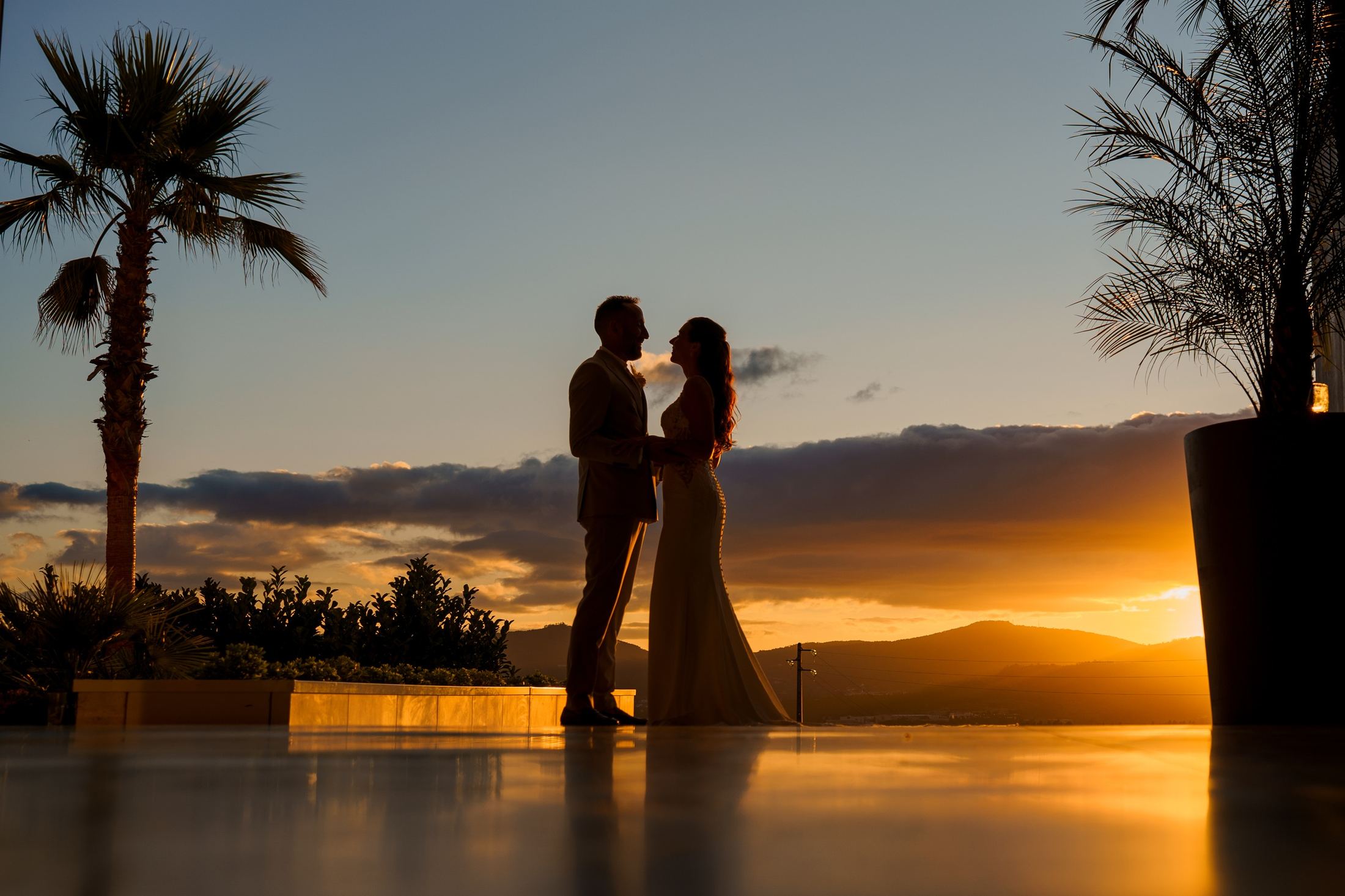 Bride and groom silhouetted at sunset beside a palm tree